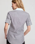 A woman with long blonde hair is seen from the back, wearing THE SHIRT's The Short Sleeve Shirt in black and white vertical stripes with black pants, against a light grey background.