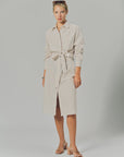 A woman wears THE SHIRT’s LS Midi Dress—a beige and white striped, tie-waist shirtdress—standing against a plain background with her hair tied back, sunglasses on her head, sandals, and a neutral expression.