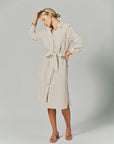 A woman poses in THE SHIRT's LS Midi Dress, a light beige cotton shirtdress with thin vertical stripes, long sleeves, a collared neckline, and a tied waist belt. She completes the look with nude sandals and her hand gently touches her forehead.