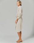 A woman stands in profile against a plain background, wearing THE SHIRT’s LS Midi Dress and tan heeled sandals, with sunglasses perched on her head.