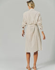 A woman with blonde hair tied back wears THE SHIRT's LS Midi Dress, a knee-length, belted cotton shirtdress with light vertical stripes, paired with light-colored heeled sandals against a plain neutral background.