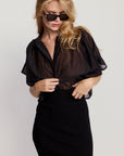 A woman in dark sunglasses wears The Hayden Shirt by THE SHIRT, a sheer black cotton voile top, paired with a high-waisted black skirt as she stands against a light background, looking down while adjusting her blouse.