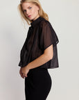 A blonde woman stands sideways against a plain light background, wearing THE SHIRT’s Hayden Shirt in sheer black cotton voile over a black skirt with a side slit. She looks at the camera with a neutral expression.