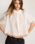 A woman with long brown hair wears THE SHIRT's cropped Hayden Shirt in lightweight cotton voile, paired with black jeans and a belt, standing hands on hips against a plain light background.