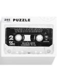 A 285-piece jigsaw puzzle box from Things Unseen featuring a vintage cassette tape design labeled "Beastie Boys Licensed to Ill." This puzzle pays homage to the iconic debut LP by highlighting detailed song titles and capturing the classic audio tape aesthetic.