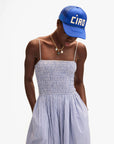 Wearing a Clare Vivier Trucker Hat Ciao, layered gold necklaces, and a light blue and white striped sundress with thin straps and a smocked bodice, the person stands with hands in pockets against a white background.