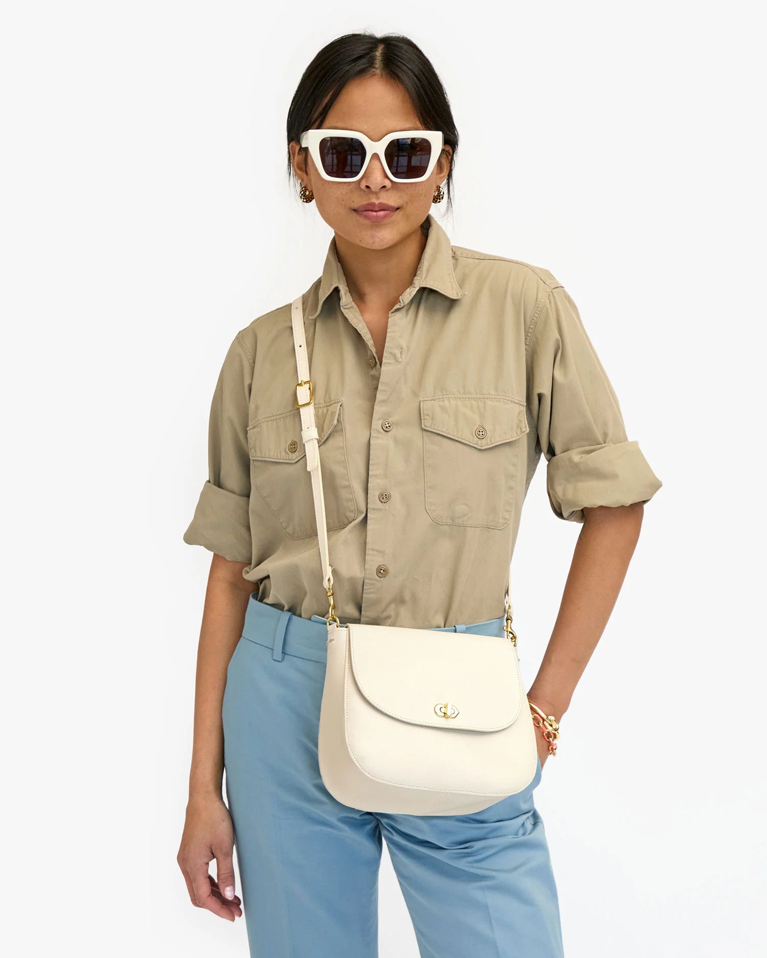 A woman wearing sunglasses, a beige shirt, and blue pants, accessorized with a Clare Vivier Turnlock Louis Cream crossbody bag holding keys and gold jewelry, standing against a plain background.