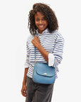 A person with curly hair smiles while holding the Clare Vivier Turnlock Louis w/ Top Handle, a sleek blue Italian leather handbag with an exterior pocket. They're in a striped long-sleeve shirt and dark jeans against a plain white background, exuding elegance.