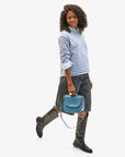 A person with curly hair wears a blue and white striped shirt, denim shorts, and knee-high black boots. They carry the Turnlock Louis w/ Top Handle by Clare Vivier, an Italian leather handbag with a crossbody strap. The background is plain white.