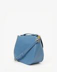 The Turnlock Louis w/ Top Handle by Clare Vivier is a stylish blue handbag made from Italian leather with a smooth finish. It features gold-toned hardware, a detachable crossbody strap, an exterior pocket, and boasts a sleek modern design against a plain white backdrop.