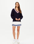 A woman with long brown hair wears The Upside's RENSHAW JOSETTE V NECK SWEATER, a navy knit with red and white trim, paired with a white pleated skirt featuring blue and red dots and white sneakers, standing against a plain white background.