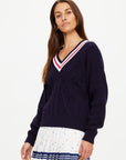 A woman with long brown hair models The Upside's RENSHAW JOSETTE V NECK SWEATER in navy blue, featuring intricate cable detailing, paired with a white pleated skirt with colorful prints against a plain light background.