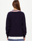 A woman with long brown hair faces away, wearing The Upside's RENSHAW JOSETTE V NECK SWEATER in navy with white, red, and navy collar detail, paired with a white pleated skirt featuring small colorful patterns.