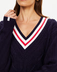 A woman wears The Upside RENSHAW JOSETTE V NECK SWEATER, an organic cotton navy blue piece with cable details and a deep V-neck accented by white and red stripes. She has light skin, long brown hair, and small gold hoop earrings.