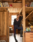 A woman in rain boots, leggings, and The Upside's RENSHAW JOSETTE V NECK SWEATER with subtle cable details stands in a cozy rustic cabin filled with books, fruit, and kitchen items, leaning casually against a wooden post.