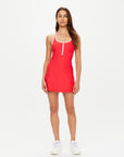 A woman stands facing forward, wearing The Upside PACIFIC ALI RACER BACK DRESS in sleeveless red with white trim, made from moisture-wicking recycled fabric, paired with white sneakers against a plain white background.