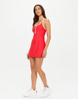 A woman with long brown hair poses confidently against a white background, wearing The Upside's PACIFIC ALI RACER BACK DRESS— a sleeveless, fitted red tennis dress with white trim made from recycled fabric—paired with white sneakers.