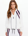 A woman with long brown hair wears The Upside's PACIFIC MORGAN ZIP UP JACKET, featuring navy blue and red stripes and raglan sleeves. She stands facing forward against a plain light background with a neutral expression.