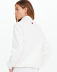 A woman with light brown hair, facing away and turning left, wears The Upside's white PACIFIC MORGAN ZIP UP JACKET featuring a small red square logo on the back, set against a plain white background.