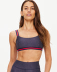 A woman with long brown hair wears The Upside's CAVALLO SCOOP NECK NINA BRA—a navy geometric print, quick-drying, mid coverage sports bra with red and navy striped straps—plus matching leggings, against a light background. Stylish and functional.