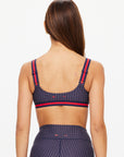 A woman with long brown hair is shown from the back, wearing The Upside's CAVALLO SCOOP NECK NINA BRA in navy with high-waisted leggings, both featuring dotted patterns, red striped accents, and made from breathable recycled fabric.