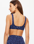 Woman with long brown hair wearing The Upside MYSTERE REVERSIBLE RORY BRA and matching leggings in space dye blue, shown from the back against a plain white background.