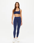 A woman faces forward in The Upside’s MYSTERE REVERSIBLE RORY BRA and matching high-waisted, space dye blue leggings with a subtle pattern and moisture-wicking fabric, styled with white sneakers against a light backdrop.