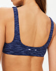 A woman shown from the back wears The Upside's MYSTERE REVERSIBLE RORY BRA in space dye blue, featuring a racerback design, moisture-wicking fabric, and a subtle white logo near the lower back.