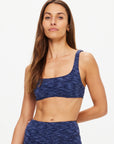 A woman with long brown hair models The Upside’s MYSTERE REVERSIBLE RORY BRA in blue, paired with matching high-waisted leggings, standing against a plain white background and looking confidently at the camera.