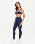 A woman wears The Upside's MYSTERE REVERSIBLE 25IN PANT—high-waisted leggings in space dye blue with a wavy pattern—paired with a matching sports bra and white sneakers, standing against a plain white background and gazing calmly to the side.