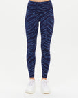 A person wears The Upside's MYSTERE REVERSIBLE 25IN PANT—high-waisted, navy leggings with a blue zebra print, made from moisture-wicking fabric—with white sneakers, standing against a plain white background.