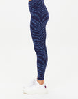 A person in profile wears The Upside MYSTERE REVERSIBLE 25IN PANT—moisture-wicking, high-waisted blue space dye leggings—with white sneakers, posed against a plain white background.