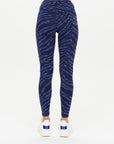 Someone wearing The Upside MYSTERE REVERSIBLE 25IN PANT—high-waisted, blue and black zebra-print, moisture-wicking, reversible recycled leggings—and white sneakers with red and blue stripes, standing with their back to a plain white background.