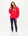 A woman with long brown hair poses against a plain background in The Upside’s RANCHERO SATURN CREW SWEATER, featuring organic cotton and the Western Logo, styled with navy leggings with red stripes and white sneakers.