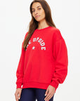 A woman with long brown hair wears The Upside's RANCHERO SATURN CREW SWEATER, a relaxed red organic cotton sweatshirt featuring a Western logo and star, paired with dark leggings, standing against a plain white background.