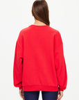 A person with straight brown hair wears The Upside's RANCHERO SATURN CREW SWEATER in bright red organic cotton, featuring a subtle puff print logo, paired with dark blue pants. They stand back to the camera against a light background.