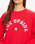 A woman wears The Upside's RANCHERO SATURN CREW SWEATER, a bright red organic cotton sweatshirt featuring a Western-style "THE UPSIDE" and star logo across the front. Only her lower face and upper torso show against a plain background.