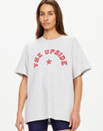 A woman with long brown hair wears The Upside's RANCHERO SAMMI TEE, an oversized organic cotton light gray top with a "THE UPSIDE" and red star Western logo, paired with dark shorts against a plain light background.