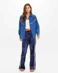 A woman faces forward against a plain white background, wearing The Upside WILDWEST SYLVE FRINGE SHIRT in blue chambray with fringe, a white crop top, patterned blue bell-bottom pants with white side stripes, and black sandals.
