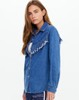 A woman with long brown hair models The Upside’s WILDWEST SYLVE FRINGE SHIRT, a western-inspired chambray top with fringe on the front and sleeves, paired with patterned navy pants. She stands slightly sideways against a plain light background.