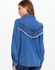 A woman facing away wears The Upside's WILDWEST SYLVE FRINGE SHIRT, a western-inspired chambray top with frayed fringe across the upper back and shoulders, paired with patterned blue pants against a plain, light background.