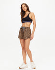 A woman with long brown hair poses confidently against a white background, wearing The Upside's PANTHERA BILLIE RUN SHORT in leopard print, a black sports bra, and white sneakers.