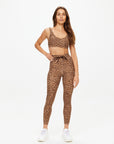 A woman with long brown hair and a relaxed expression stands against a white background wearing The Upside's PANTHERA 25IN MIDI PANT and matching leopard print sports bra, paired with white sneakers and one hand on her hip.