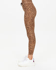 A person stands sideways against a plain white background, wearing The Upside’s PANTHERA 25IN MIDI PANT—high-waisted, moisture-wicking leopard print leggings—paired with white sneakers.