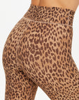 A close-up side view of a person wearing The Upside PANTHERA 25IN MIDI PANT, featuring moisture-wicking brown and black leopard print leggings, against a plain light background.