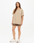A woman with long brown hair and a relaxed expression stands against a white background wearing The Upside's PALOMINO SAMMI TEE in oversized tan, paired with leopard print shorts and white sneakers.