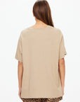 A woman stands facing away from the camera, wearing The Upside's PALOMINO SAMMI TEE in a neutral tan shade with leopard print shorts, set against a plain white background.