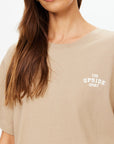 A woman wears The Upside’s PALOMINO SAMMI TEE, an oversized neutral tan t-shirt with "THE UPSIDE SPORT" in white on the chest. Only her lower face, neck, and upper torso are visible.