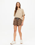 A woman with long brown hair poses casually against a white background, wearing The Upside's PALOMINO SAMMI TEE in a neutral tan shade, paired with leopard print shorts and white sneakers.
