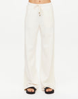 A model wears The Upside BOW N ARROW NOVA FLARE PANT—loose-fitting, cream-colored wide leg pants—paired with black platform sandals, shown from the waist down against a white background.